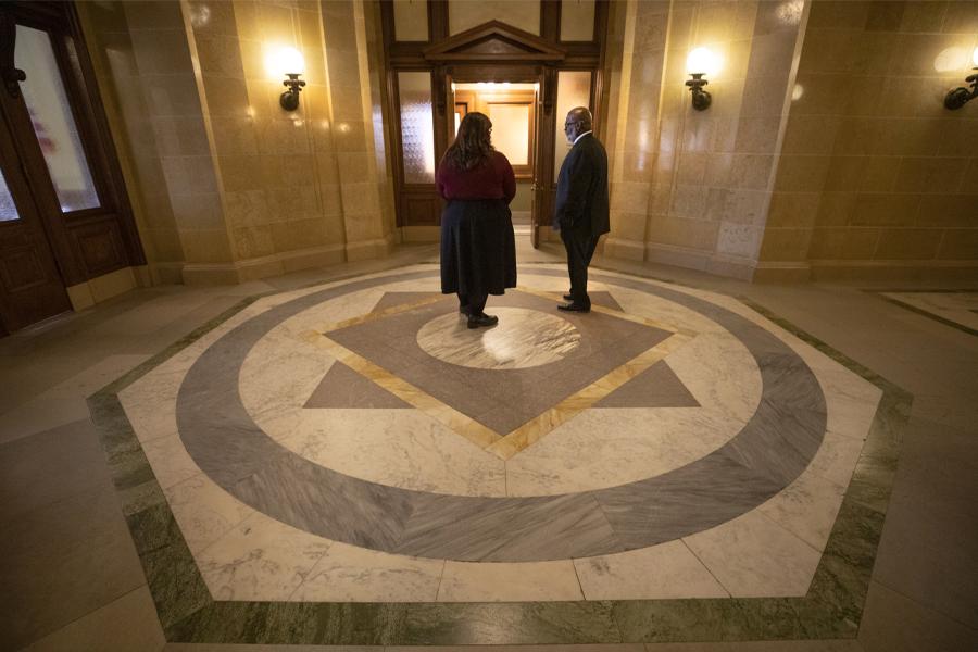 Two people talk in a room with dark woodwork and American flags.